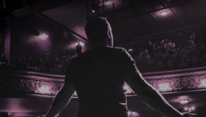 Highly stylised shot of Keith performing in front of a theatre audience, his back to the camera and arms outstretched